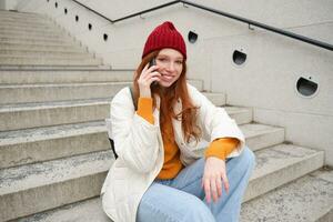 joven elegante pelirrojo niña en rojo sombrero, se sienta en calle y negociaciones en móvil teléfono, tiene teléfono conversación, anillos su amigo mientras relaja al aire libre foto