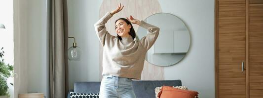 Women and wellbeing. Beautiful young asian woman dancing, feeling carefree and happy, raising her hands above head and smiling, enjoying her stay at home photo