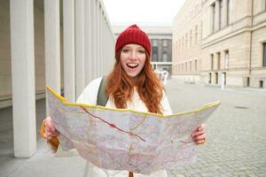 hermosa pelirrojo mujer, turista con ciudad mapa, explora Turismo histórico punto de referencia, caminando alrededor antiguo ciudad, sonriente felizmente foto