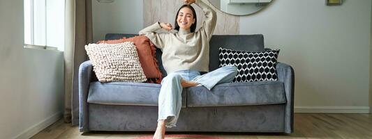 Portrait of happy asian woman feeling lazy, stretching on sofa and smiling pleased, relaxing at home, resting from work photo