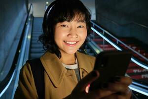cerca arriba de linda asiático niña en auriculares, escoge canción a escucha mientras viajando, yendo abajo escalera mecánica a metro, sonriente como leyendo mensaje en móvil teléfono foto