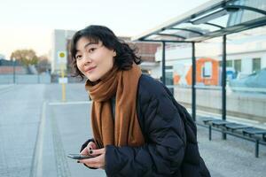 linda sonriente mujer en pie en un autobús detener y mirando a camino, esperando para su público transporte, participación teléfono inteligente, comprobación calendario en móvil aplicación foto