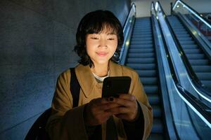 Happy smiling young woman, standing on escalator, going down, holding smartphone in both hands, chatting on mobile phone app photo