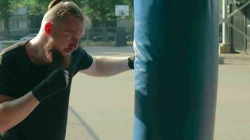 Street Fighter in Black Clothes and Bandages on the Wrist Boxing in Punching Bag Outdoors. Young Man Doing Box Training and Practicing His Punches at the Outside Gym Slow Motion video