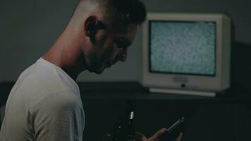 Man in White Tshirt is Sitting Against the Old Silver TV in Dark Room. Drinking Beer and Using His Smartphone. Search for TV Signal. Distortion on the Screen. Back VIew, Medium CloseUp, Film Grain video