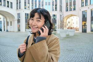 celular tecnología. joven coreano mujer negociaciones en móvil teléfono, hace un teléfono llamada en su camino hogar, camina abajo calle, ciudad centro, tiene teléfono conversacion foto