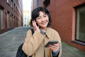 retrato de joven coreano mujer caminando alrededor ciudad con mochila y auriculares, escucha música, mira a teléfono inteligente, usos teléfono solicitud en calles foto