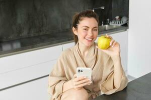 Close up portrait of smiling brunette woman in bathrobe, sits in kitchen at home, uses mobile phone and holds an apple, orders fresh fruits on smartphone app, buys groceries online, looks up recipe photo