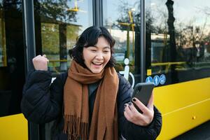 Portrait of cheerful asian girl talks on mobile phone, video chats, looks amazed at smartphone camera, stands on bus stop photo