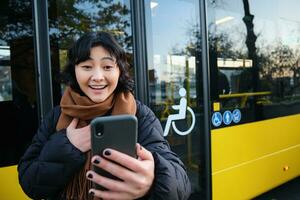 entusiasta asiático mujer, en pie en autobús detener con teléfono inteligente, mirando a teléfono pantalla con asombrado, triunfando rostro, victorioso, oír genial Noticias en vídeo charla foto