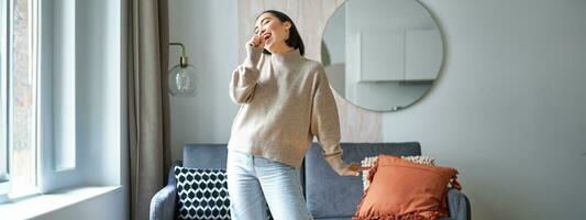 People and emotions. Portrait of asian girl standing in living room, singing in shadow microphone, dancing and having fun, feeling joy photo