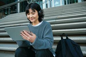 Young asian girl, student in headphones, works on remote, digital artist drawing on tablet with graphic pen, listening music in headphones and sitting on street staircase photo