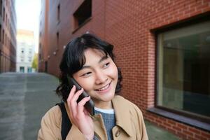 celular tecnología concepto. joven moderno niña negociaciones en móvil teléfono, camina en calle y sonrisas, tiene conversacion terminado teléfono foto