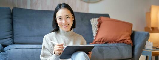 Portrait of asian woman with tablet, drawing, working on design project, holding pen, sitting in her living room, freelancing photo