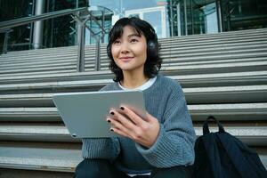 Young asian girl, student in headphones, works on remote, digital artist drawing on tablet with graphic pen, listening music in headphones and sitting on street staircase photo