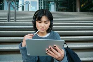 Portrait of asian girl with troubled face, draws on digital tablet with pen, looks complicated at her work, feels stuck no inspiration, sits on stairs and thinks photo