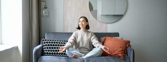 relajación y paciencia. sonriente joven asiático mujer en acogedor habitación, sentado en sofá y meditando, haciendo yoga atención plena formación foto