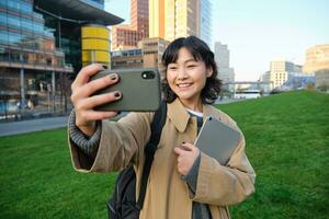 contento Universidad alumno, coreano niña toma selfie con su documentos y digital tableta, sostiene teléfono inteligente y poses cerca Universidad instalaciones foto