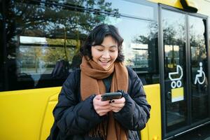 Beautiful korean girl, student on bus stop, looking at her smartphone, checking timetable, reading text message, wearing winter clothes photo