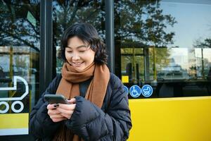 Young beautiful woman standing on bus stop, texting message on smartphone, holding mobile phone, checking her schedule, buying ticket online, wearing winter clothes photo