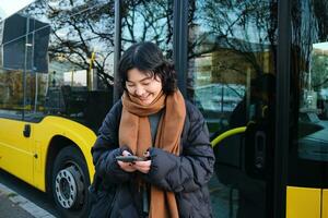 Beautiful korean girl, student on bus stop, looking at her smartphone, checking timetable, reading text message, wearing winter clothes photo