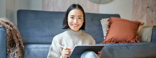 Portrait of asian woman with tablet, drawing, working on design project, holding pen, sitting in her living room, freelancing photo