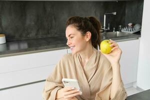 Image of attractive young woman in bathrobe, sits in kitchen, eats green apple and looks at mobile phone, uses smartphone app, orders food on application photo