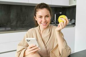 Close up portrait of happy young woman in bathrobe, sitting in the kitchen and using mobile phone, holding an apple, order fruits and vegetables online, using smartphone app for groceries delivery photo