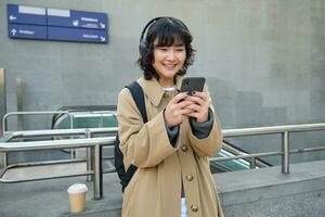 Portrait of stylish asian hipster girl, listens music in headphones, looks at her phone, tourist looking at map for a way to sightseeing, drinking coffee to go photo