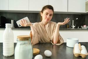 Attractive young cheerful girl baking at the kitchen, making dough, holding recipe book, having ideas photo