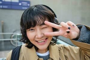 cabeza retrato de coreano niña en pie en calle, demostración paz firmar, vistiendo auriculares, escuchando música y sonriente foto