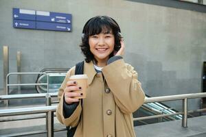 Beautiful smiling Korean girl, drinks takeaway coffee, listens music in headphones, wears trench and backpack, stands on train station, travelling photo