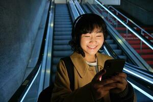 retrato de niña estudiante en auriculares, escucha música, viajes diarios, va abajo escalera mecánica, mira a teléfono inteligente con satisfecho sonrisa, contento cara foto