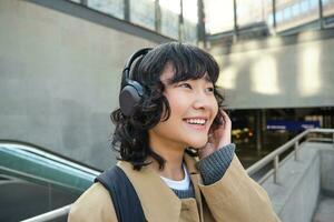 cerca arriba retrato de coreano niña en auriculares, estudiante escucha música en auriculares y sonrisas, camina alrededor ciudad, turista de viaje foto