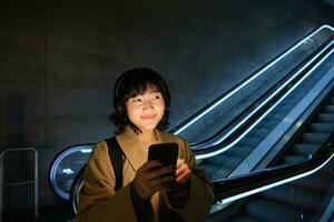 Smiling Korean girl in headphones, listens music, uses mobile phone in tube, stands near escalator in dark, looks cheerful photo