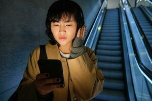 Woman in headphones, listens music, looks concerned at her phone screen, goes down escalator in city photo