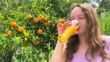 de flicka drycker orange juice mot de bakgrund av en mandarin träd, den kan vara orange juice mandarin mango hon drycker girigt och verkligen gillar de juice utsökt överallt grönt och sommar video