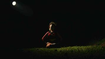 an Asian woman is sitting on a green grass while dancing with her hands and a red scarf in a forest video