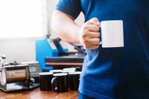 unrecognizable man holding awhite cup behind a bottom where there are cups and adesign sublimation machines. photo