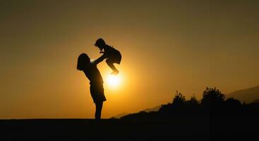 silueta, contento madre y hija abrazo lanzar alto en el campo césped a puesta de sol en el noche. amor familia actividad relajarse concepto. foto