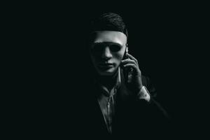 Unknown businessman wearing mask with covered face using mobile phone makes an anonymous call intimidating and threatening the interlocutor on dark background. hacker callcenter concept. photo