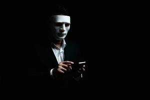 Unknown businessman wearing mask with covered face using mobile phone makes an anonymous call intimidating and threatening the interlocutor on dark background. hacker callcenter concept. photo