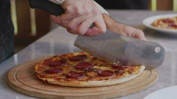 ein Mann im ein Schürze zieht Pizza aus von ein holzbefeuert Ofen. ein Koch zieht aus ein gekocht Pizza. Verbrennung Essen im ein holzbefeuert Ofen. groß Messer schneidet Pizza mit Käse. Kochen Essen Über ein öffnen Feuer. natürlich video