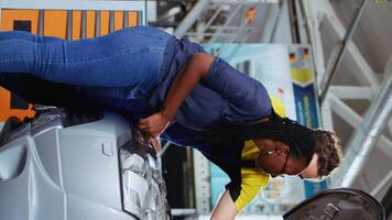 vertical vídeo trabajo en equipo colegas en garaje espacio de trabajo fijación coche para clientes, a fondo comprobación debajo vehículo capucha. mecánica haciendo rutina mantenimiento en automóvil en auto reparar tienda video