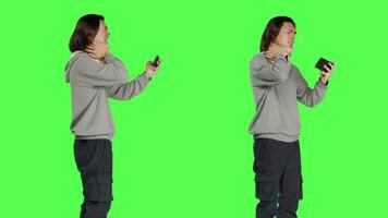 Asian man filming video on phone, wearing cervical neck brace over greenscreen in studio. Young adult recording montages or shots while he deals with bone fracture using foam collar brace.