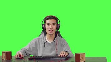 POV of adult enjoying video games on computer while speaking on headset to his colleagues, playing interactive combat contest at office desk. Asian gamer having fun with leisure time.