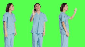 Medical specialist presents okay gesture over greenscreen backdrop, showing approval and like symbol. Healthcare worker in uniform doing agreement sign standing in studio. video