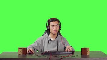 POV of asian adult enjoying computer games at desk while sitting over full body greenscreen backdrop. Gamer has fun with friends online in gaming web challenge, using headphones at desktop. video