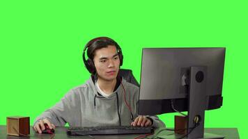 asiatico giocatore gioco su computer a scrivania, giocando in linea videogiochi con le persone. giovane adulto avendo divertimento con rpg concorrenza, A proposito di per amici su cuffie al di sopra di schermo verde. video