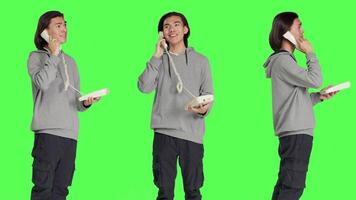 Asian person talking on landline phone, answering call on telephone with cord while he stands against greenscreen backdrop. Young adult using retro phone for remote communication. video